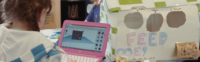 a child designing on a laptop, a cardboard model beside her